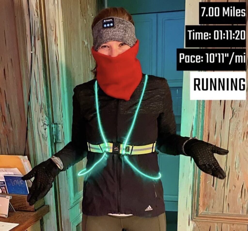 A woman is standing inside wearing a black jacket, black gloves, red neck warmer, and grey ear band. She has a reflective vest on that is glowing green. There is text in the top right corner saying she ran 7 miles in a time of 1 hour, 11 minutes, 20 seconds, with an average pace of 10 minutes 11 seconds per mile.