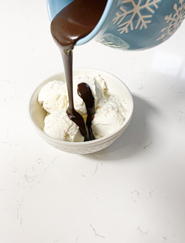 A light blue pitcher with white snowflakes is pouring hot fudge over a white bowl of vanilla ice cream.