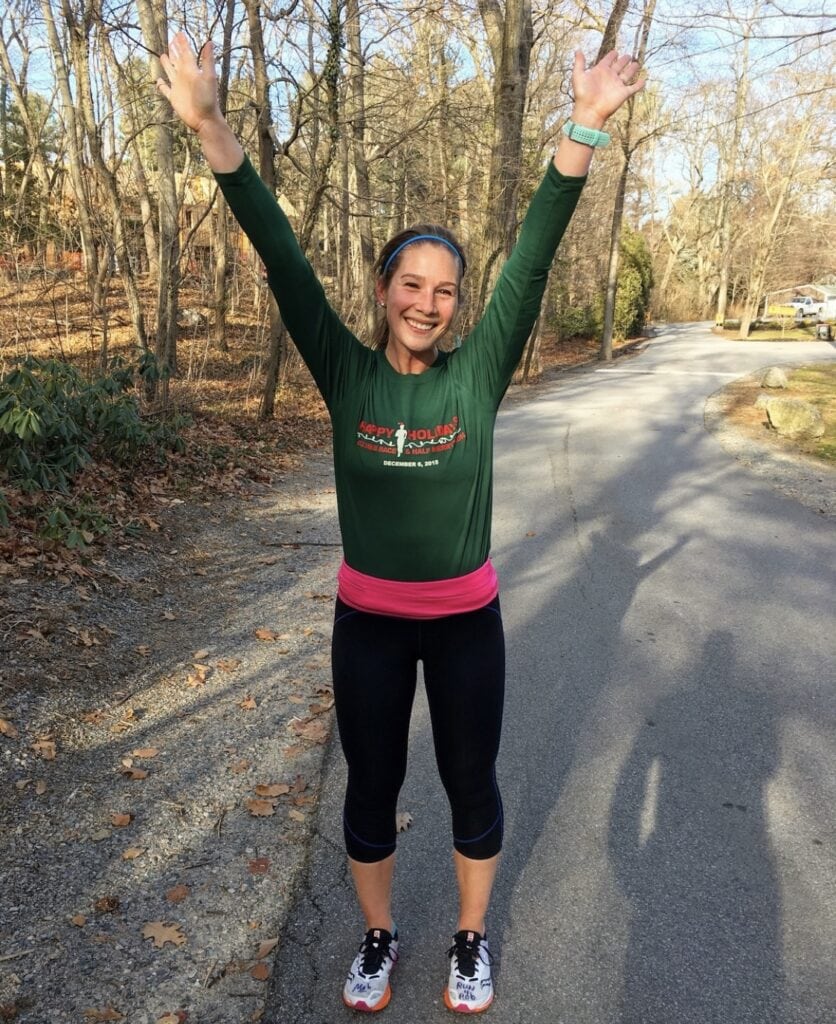 Woman in a green long sleeve shirt and black kapri leggings is standing outside on a driveway. She is looking at the camera and smiling with her arms stretched above her head.