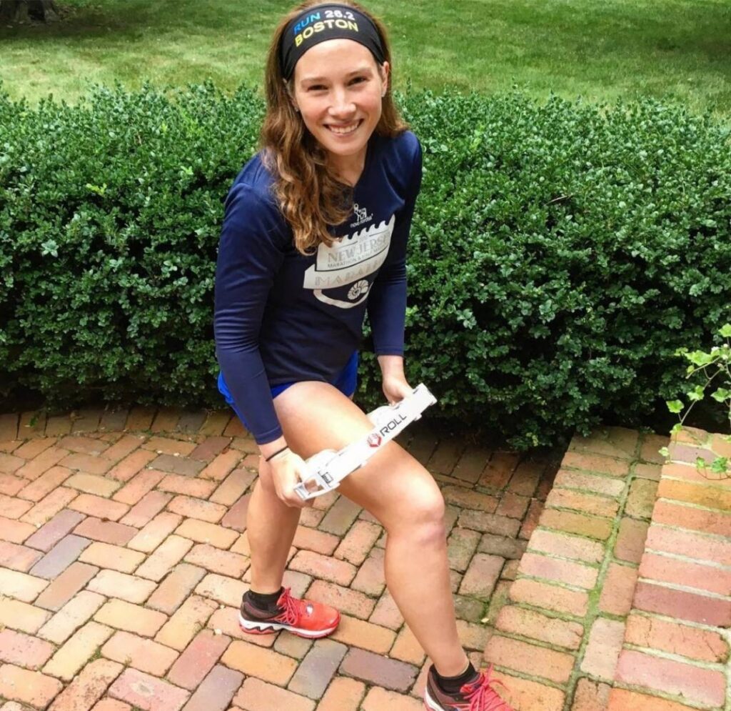 A woman is standing outside with one foot up on a step. She is massaging her right leg with a roll recovery device. She is wearing a black headband and a navy blue long sleeve shirt. Her hair is down and she is looking at the camera and smiling. She is focusing on recovery as recovery is one guideline in how to return to running after time off.  