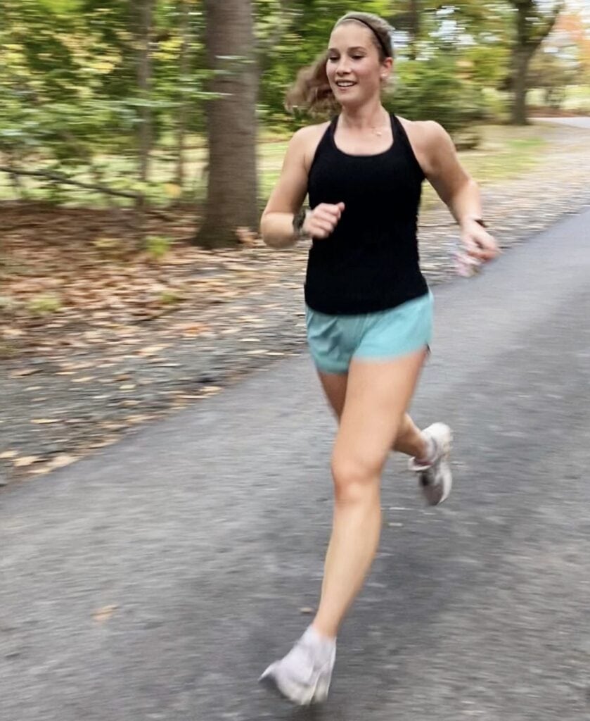 A woman is running down a driveway. She is wearing a black tank top and sea green shorts. She is smiling. 