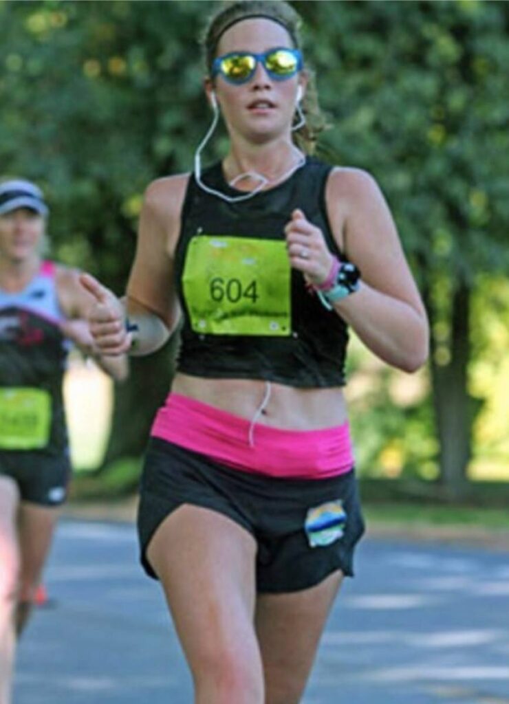 A woman is running in a road race. She is wearing a black shirt, black shorts, and headphones. She has a bright pink running belt around her waist. 