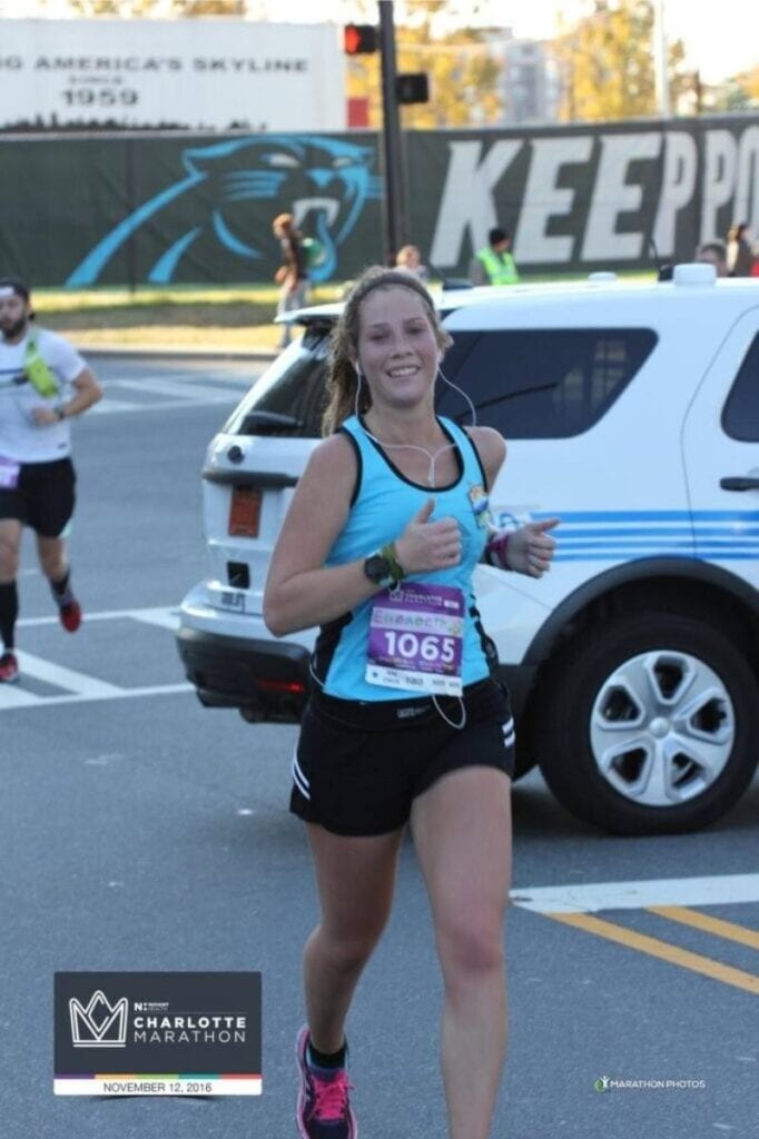 A woman is running a marathon. She is wearing a blue tank top and black shorts. She is smiling at the camera and giving two thumbs up.