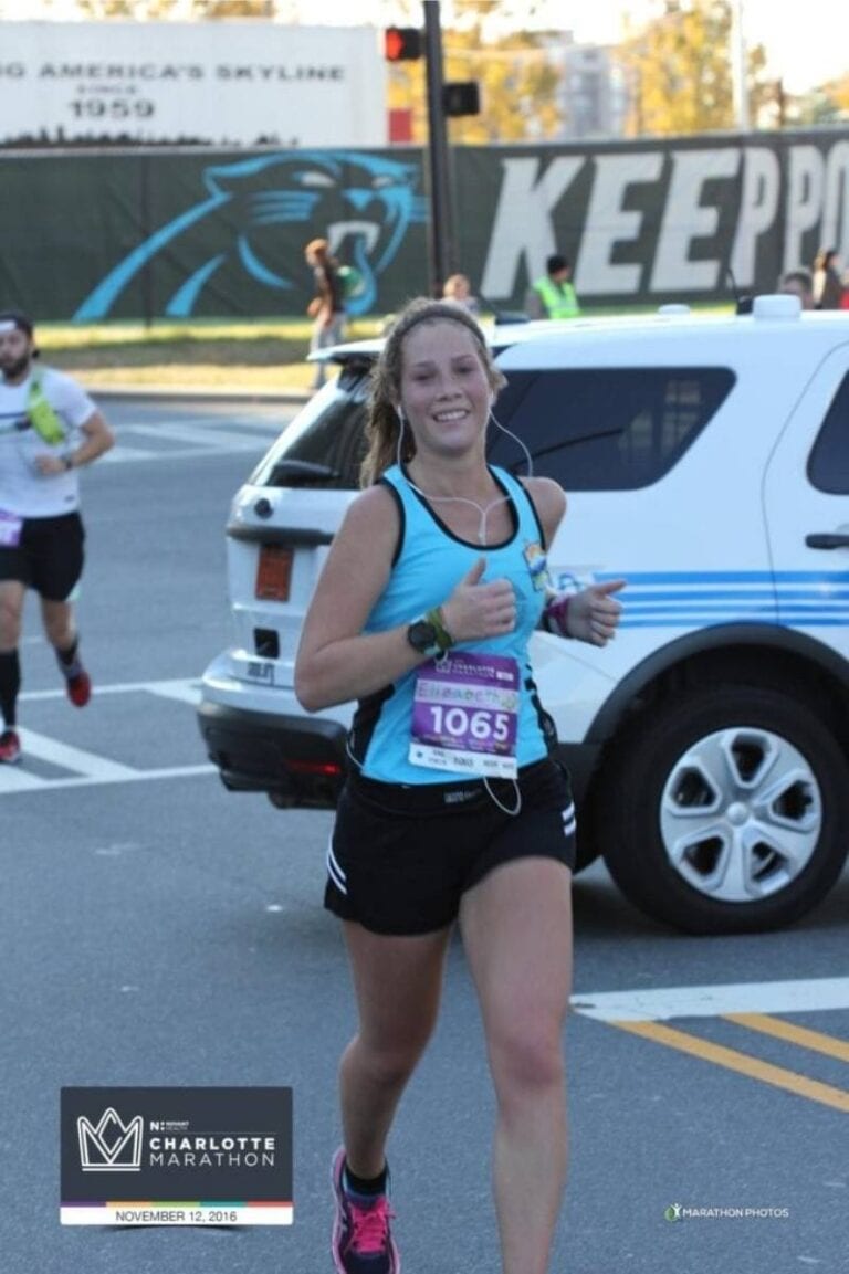 A woman is running a marathon. She is wearing a blue tank top and black shorts. She is looking at the camera while she's running and giving two thumbs up. Having fun and smiling, like this woman is, is one way to run your best race.