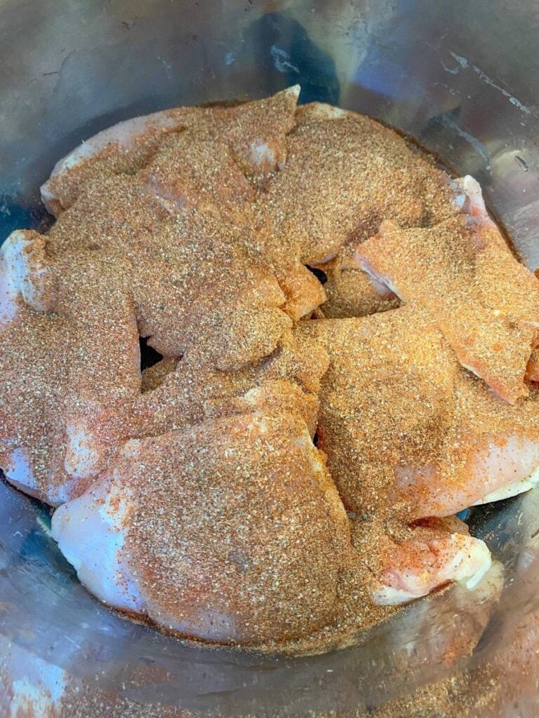There are raw chicken thighs in a metal pot with seasonings sprinkled on top.