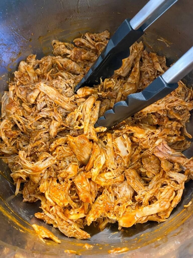 This is a closeup photo of Shredded BBQ Chicken in a metal pot. There is a pair of tongs in the chicken.