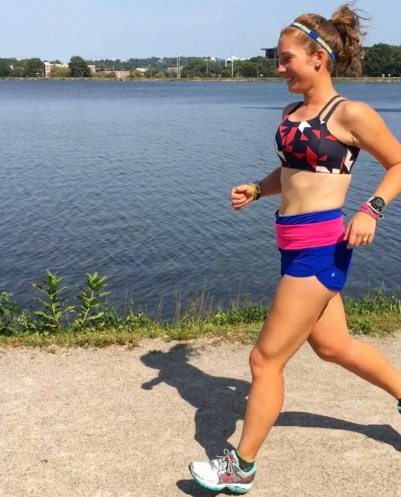 A woman is running on a dirt path around a body of water. She is wearing blue shorts, a pink waistband, and a red and navy blue sports bra. She is smiling. 