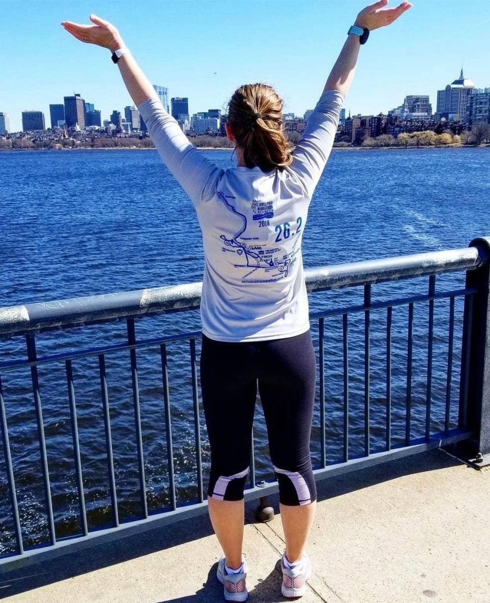 A woman is wearing a grey long sleeve shirt and black leggings. She is standing on a bridge looking out over a large body of water. We are looking at her back and she is holding her arms in the air.