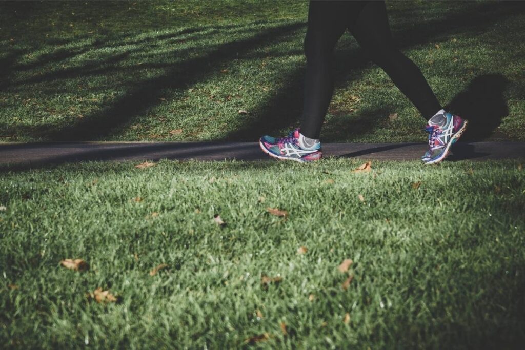A person is outside on a path walking. We can only see their legs and feet. The person is wearing black leggings, white socks, and running shoes. The is grass surrounding the path. Recovery for runners begins during the cool down. 