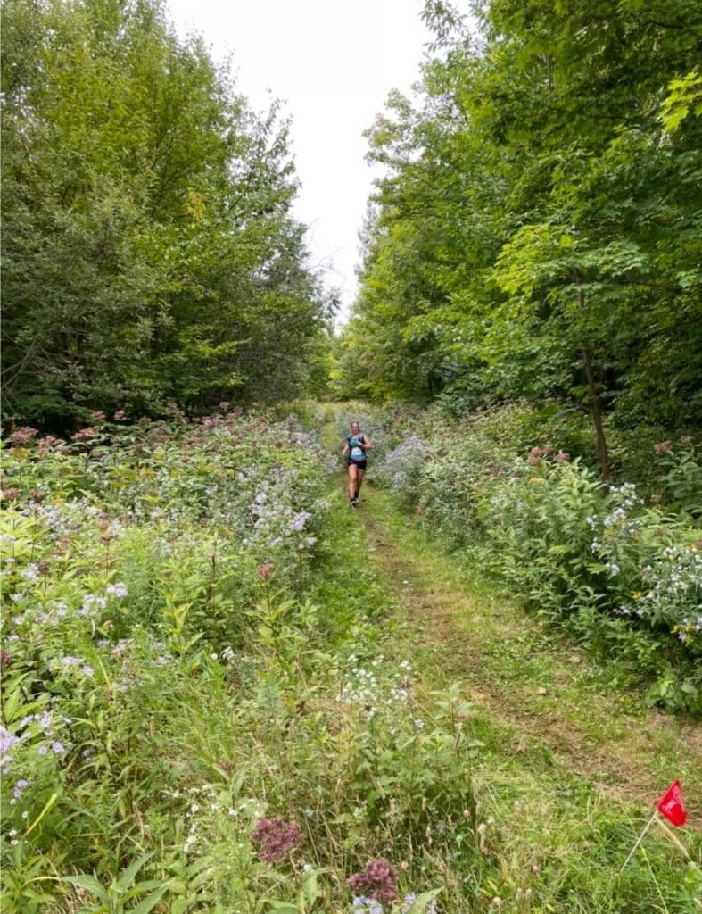 A woman is running down a lush green mountain. She is on a single track trail. She is wearing a blue tank top and black shorts. 