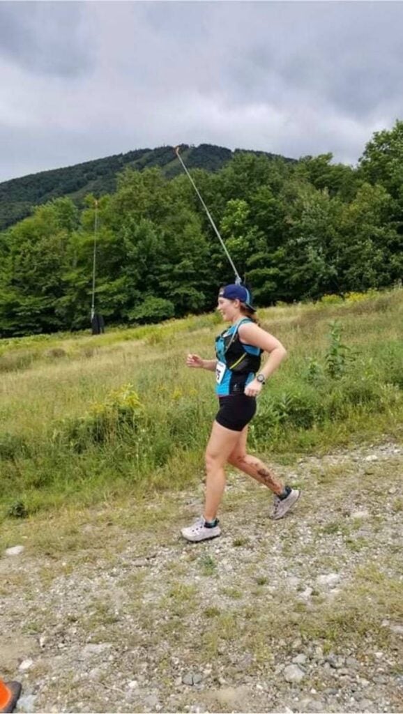 A woman wearing a blue tank top and black shorts is running a trail race. She is running down a trail wearing a black hydration vest and a backwards hat. 