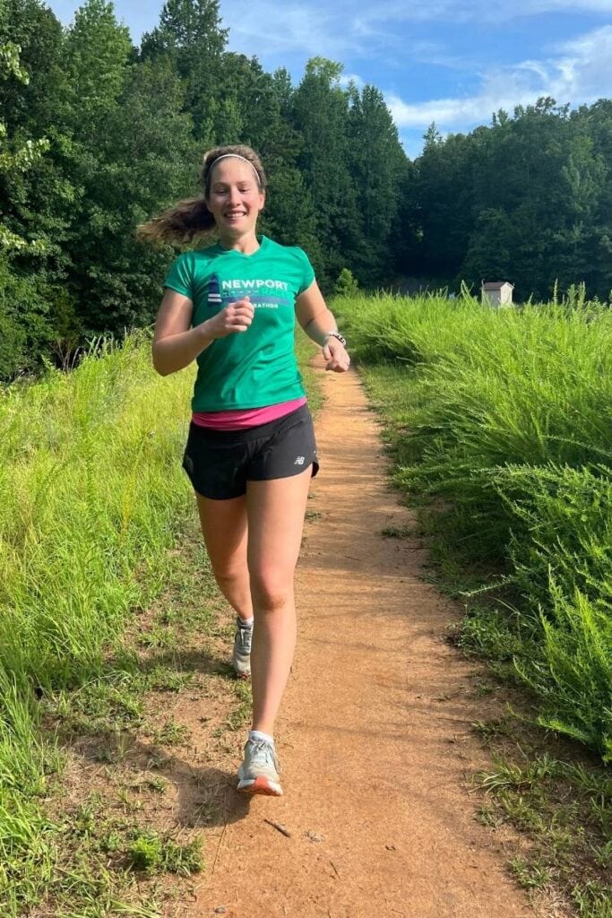 A woman is wearing a green t-shirt, black shorts, and a pink running belt. She is running on a dirt path on a trail. The woman is looking down and smiling. Running on trails is one way to change up your running routine while maintenance running. 