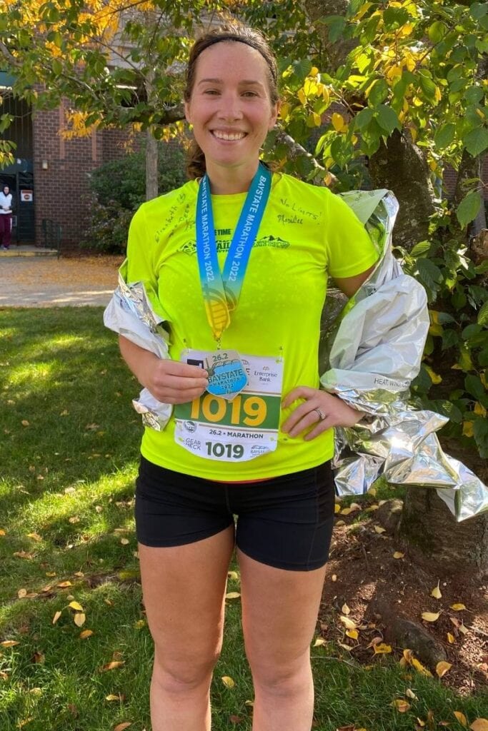 A woman wearing a bright yellow t-shirt and black shorts is standing in front of a tree. She is wearing a marathon medal around her neck and is smiling at the camera. 