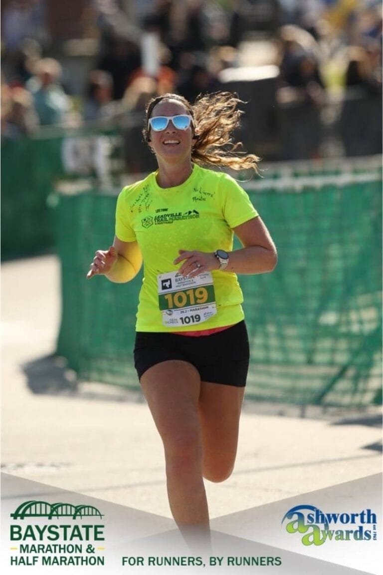 A woman wearing a bright yellow t-shirt, black shorts, and white sunglasses is approaching the finish line of a marathon. She is running and smiling.