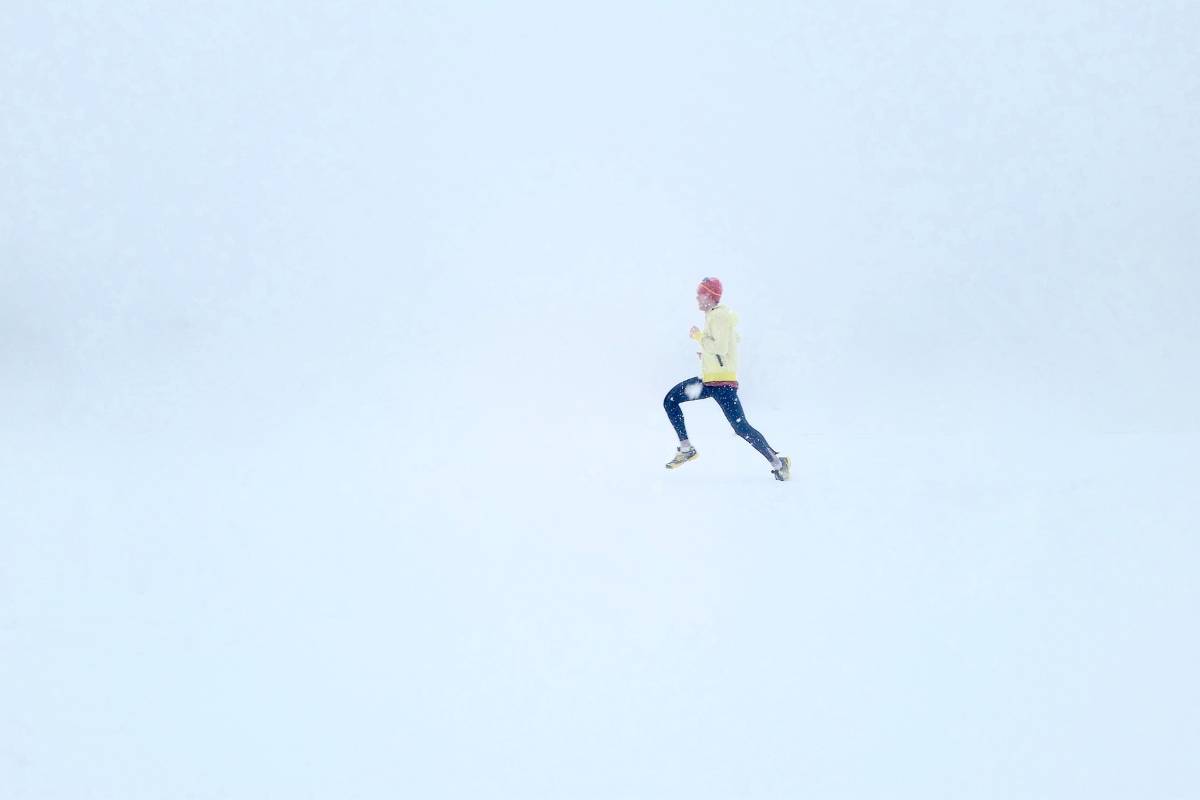 A man is running on snow with lots of fog and snow around him. He is wearing a red hat, yellow jacket, and navy blue leggings.