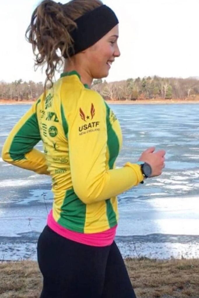 A woman is running in front of a body of water in winter. She is wearing a yellow and green long sleeve shirt with a black headband. RPE running is something to focus on during winter training. 