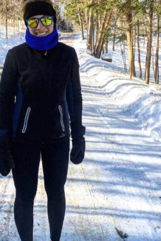 A woman is standing on a snow covered road. She is wearing a black jacket, leggings, headband, and mittens with blue sunglasses and a neck warmer. She is smiling at the camera. Not wearing the appropriate amount of layers is a common winter running mistake. 