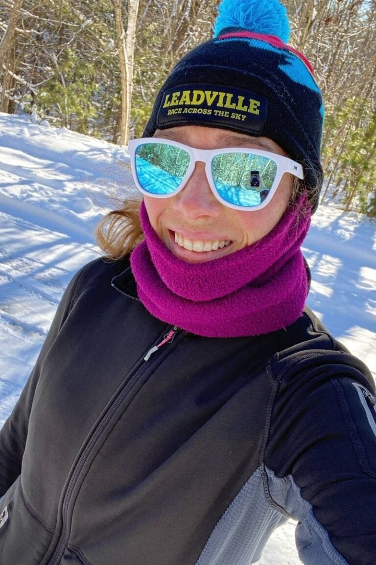 A woman is outside on a snowy road and taking a selfie. She is wearing a black hat, white sunglasses, purple neck warmer, and black jacket. Dressing warmly and wearing eye protection is ideal for what to wear for cold weather running.