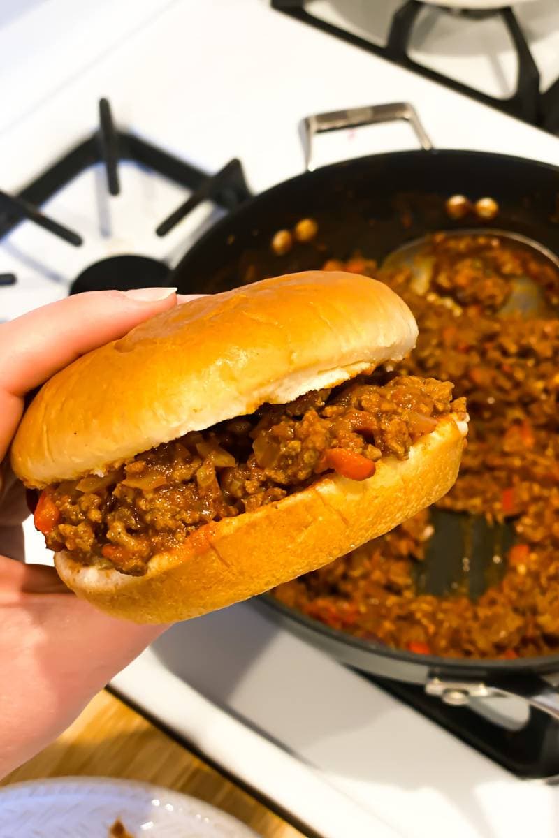 Someone is holding a hamburger bun with simple sloppy joes in it above a stovetop. There is a pan in the background that has more simple sloppy joes in it.