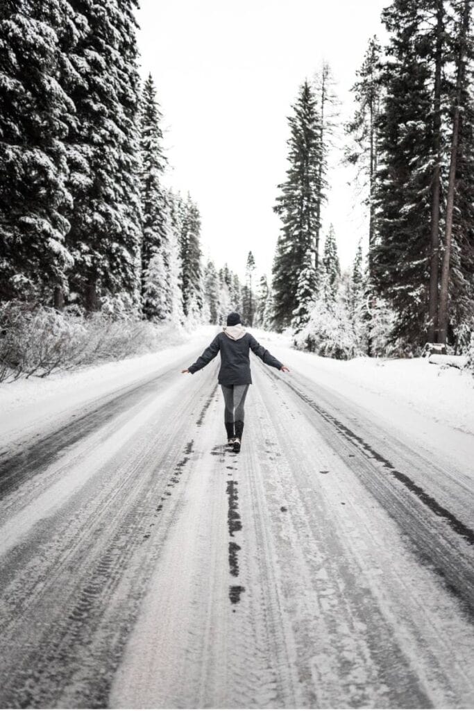 This is a black and white photo of a female walking down a snow covered road. She is in the middle of the road and her arms are out to her side like she is trying to balance. She is walking away from the camera. 