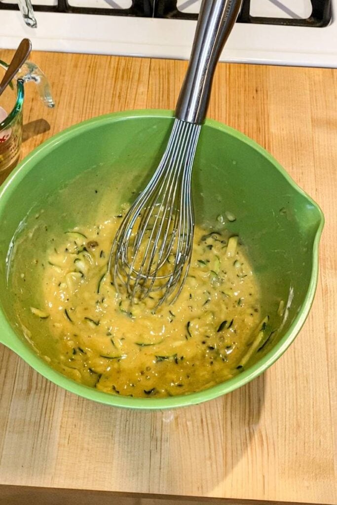 The wet ingredients for the bread are combined in a green mixing bowl. There is a silver whisk lying inside the bowl. 