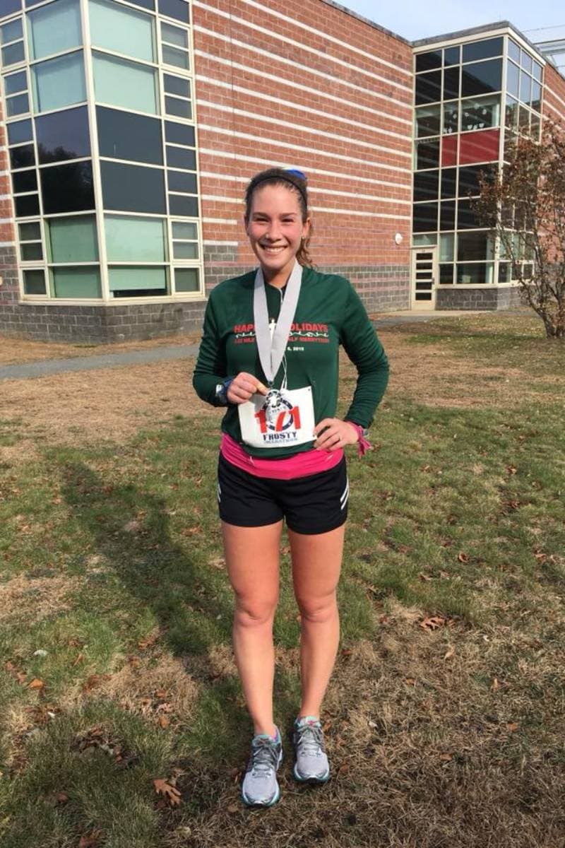 A woman is standing on the grass in front of a building. She has on a green long sleeve shirt and black shorts. She is wearing a race medal around her neck and smiling. Celebrating your achievement is a part of race recovery.
